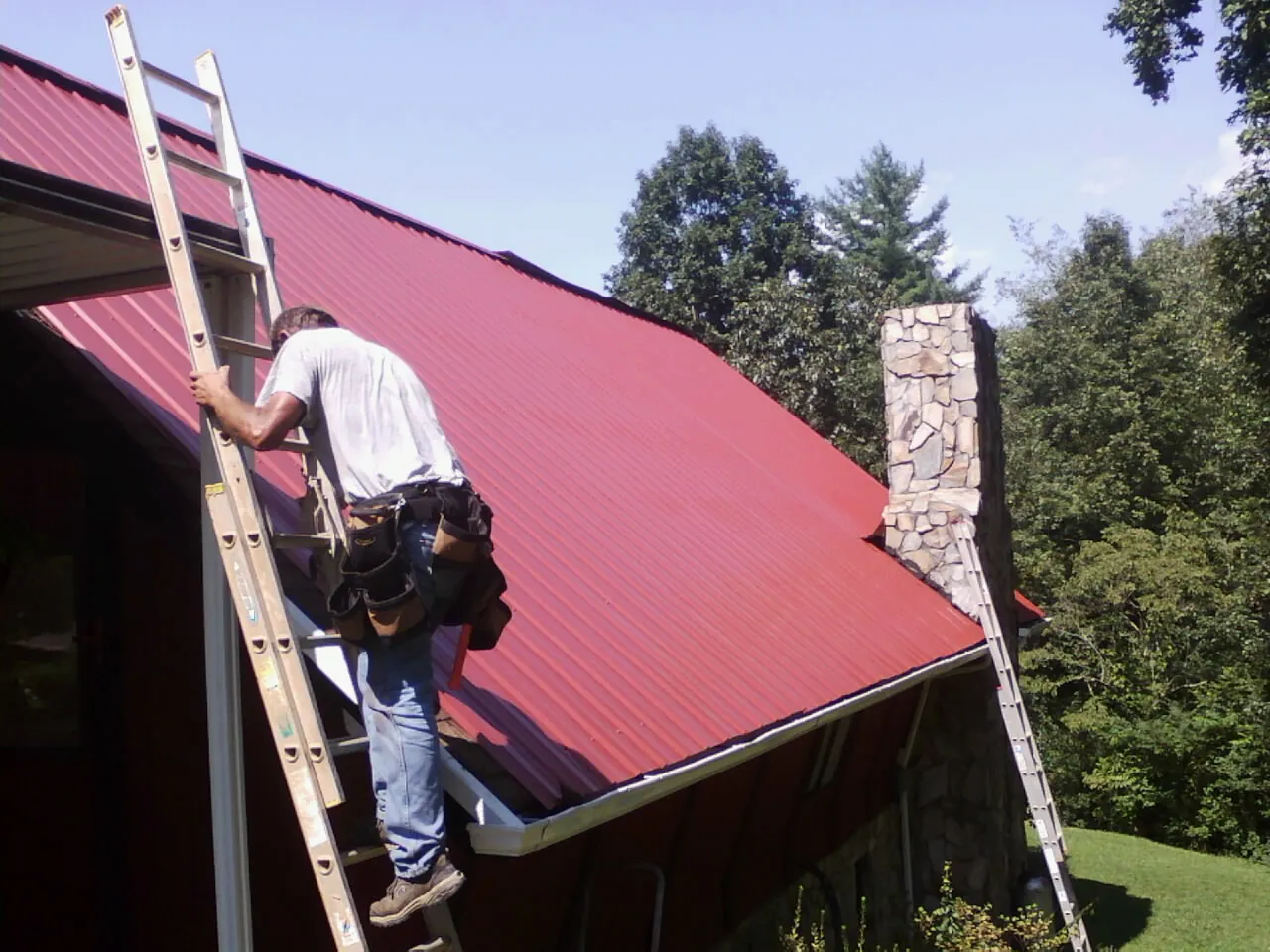 Roofer on a ladder beside a red metal roof and stone chimney — Soffit & Fascia Repair by Roofing Brothers in Bowers