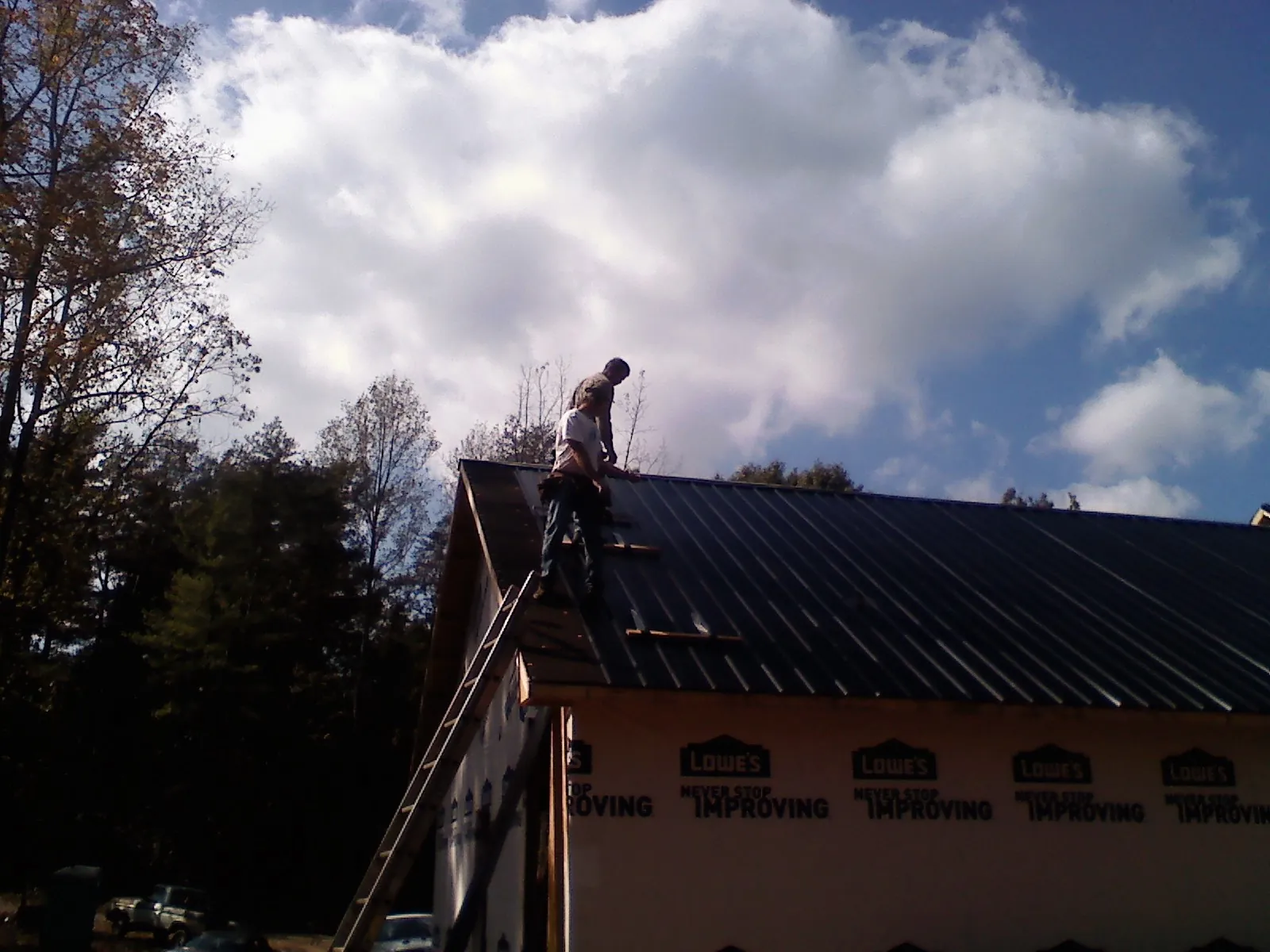 Roofer fastening dark metal panels high against a cloudy sky, from a Roof Inspection job in Bowers